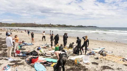 A group of surfers and friends in wetsuits gathering gear on a sandy beach, waves and a rocky point in the background.