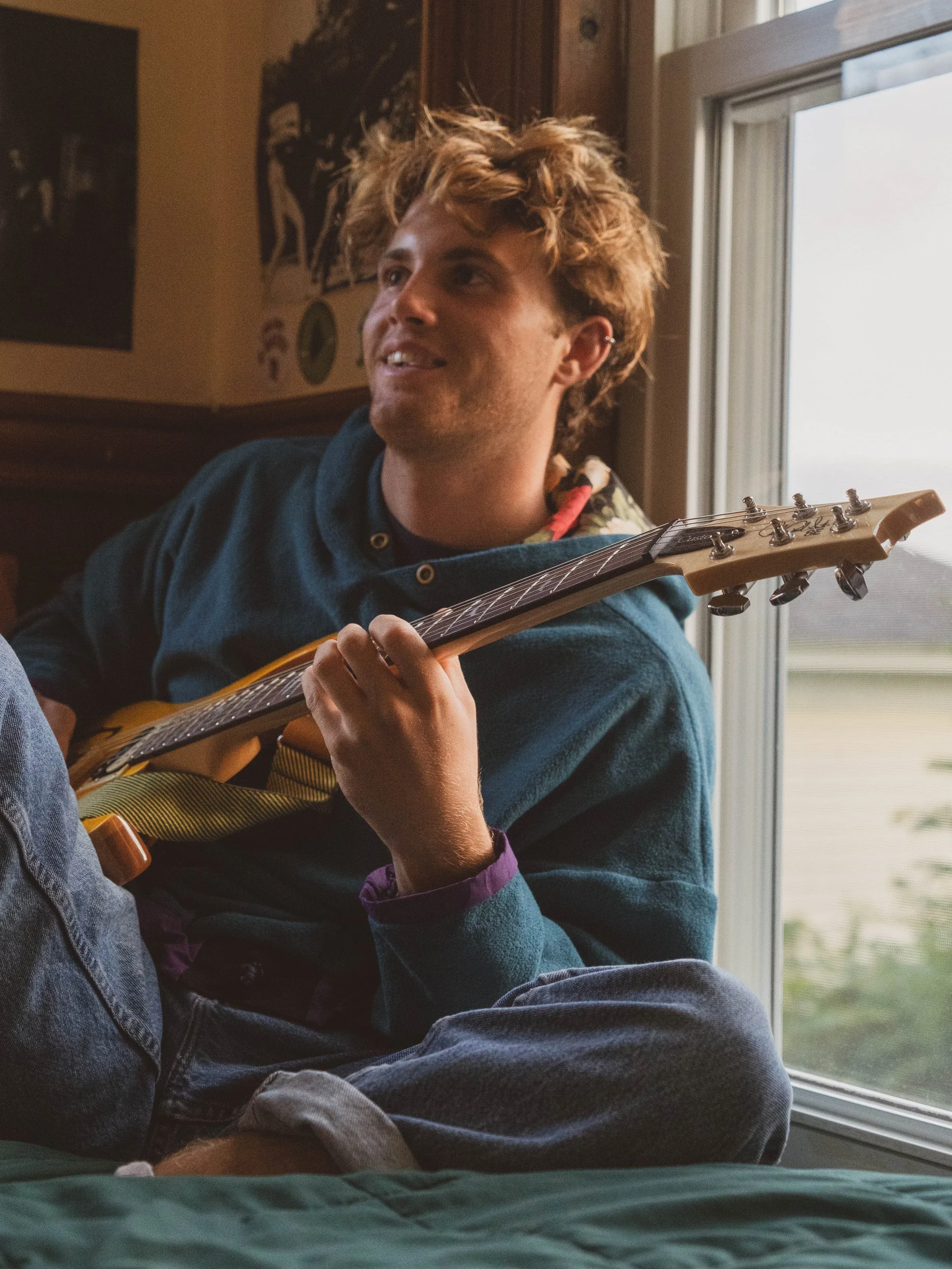 A young man playing an electric guitar and smiling, looking off to the side, near a sunlit window.