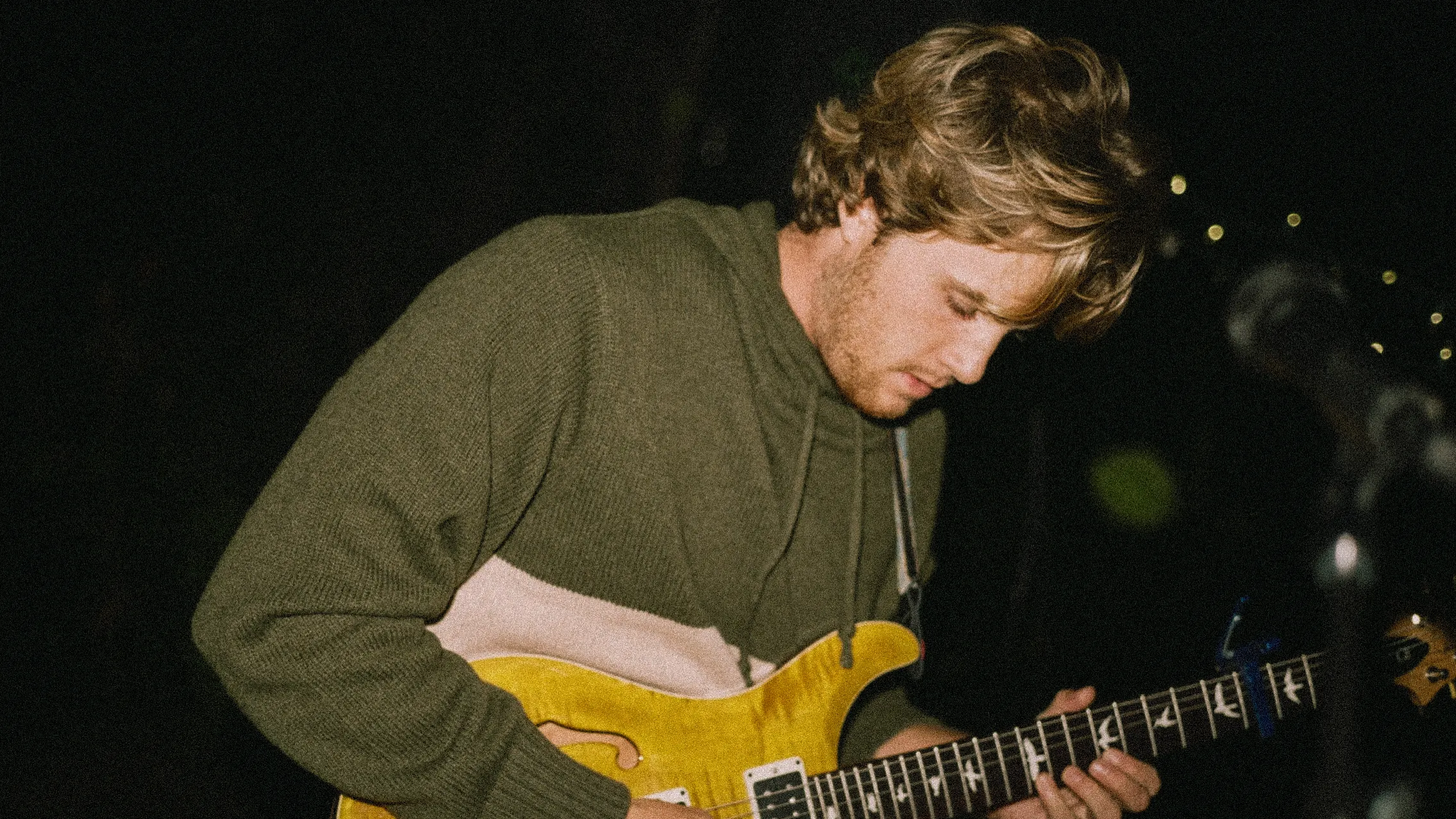 Eno Little playing a yellow-finish PRS electric guitar on a dark stage, wearing an olive green hooded sweater, warm stage light on his face and hair, soft bokeh lights in the background, film grain.