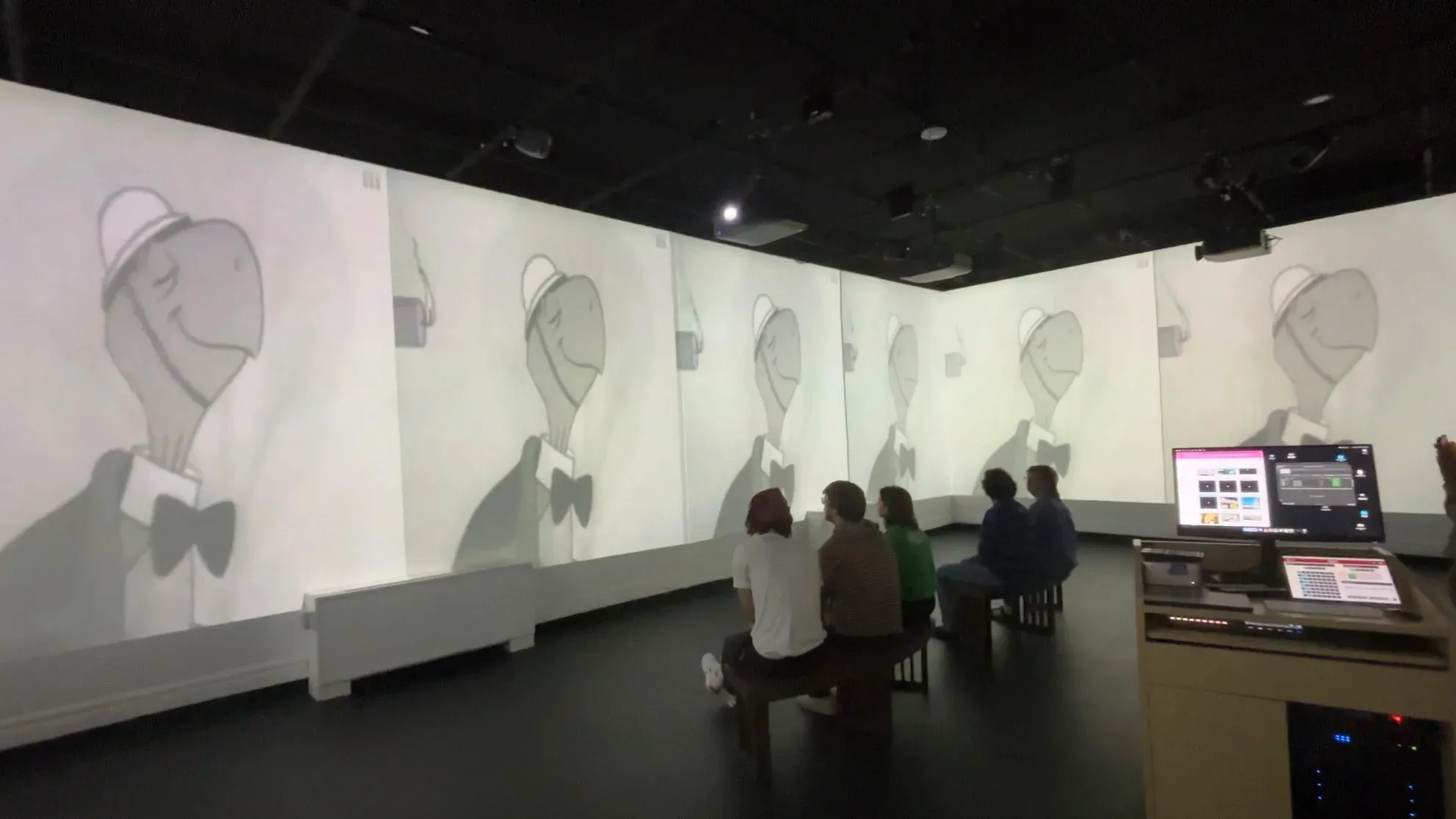 Installation view of a cartoon "friendly atom" mascot animation projected across a wraparound screen, viewers seated on benches below.