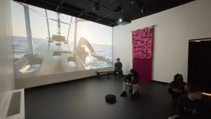 Installation view of a two-screen projection showing footage from a sailboat at sea, viewers seated on a bench beside a pink woven textile hanging on the wall.