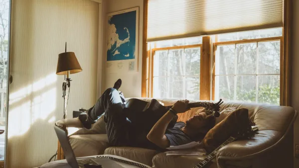 A man lying on a leather couch playing an acoustic guitar with his feet up, sunlight streaming through the windows.
