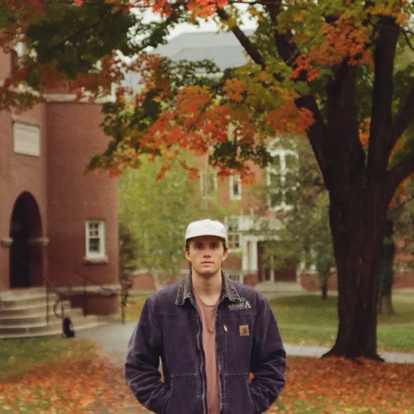 A young man in a cap and denim jacket standing on a New England college campus quad under orange autumn foliage.