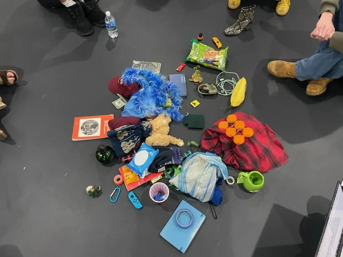 Overhead view of a pile of personal items — a stuffed toy, ball caps, snack bags, a book — collected on the floor, participants' feet visible around the edges.