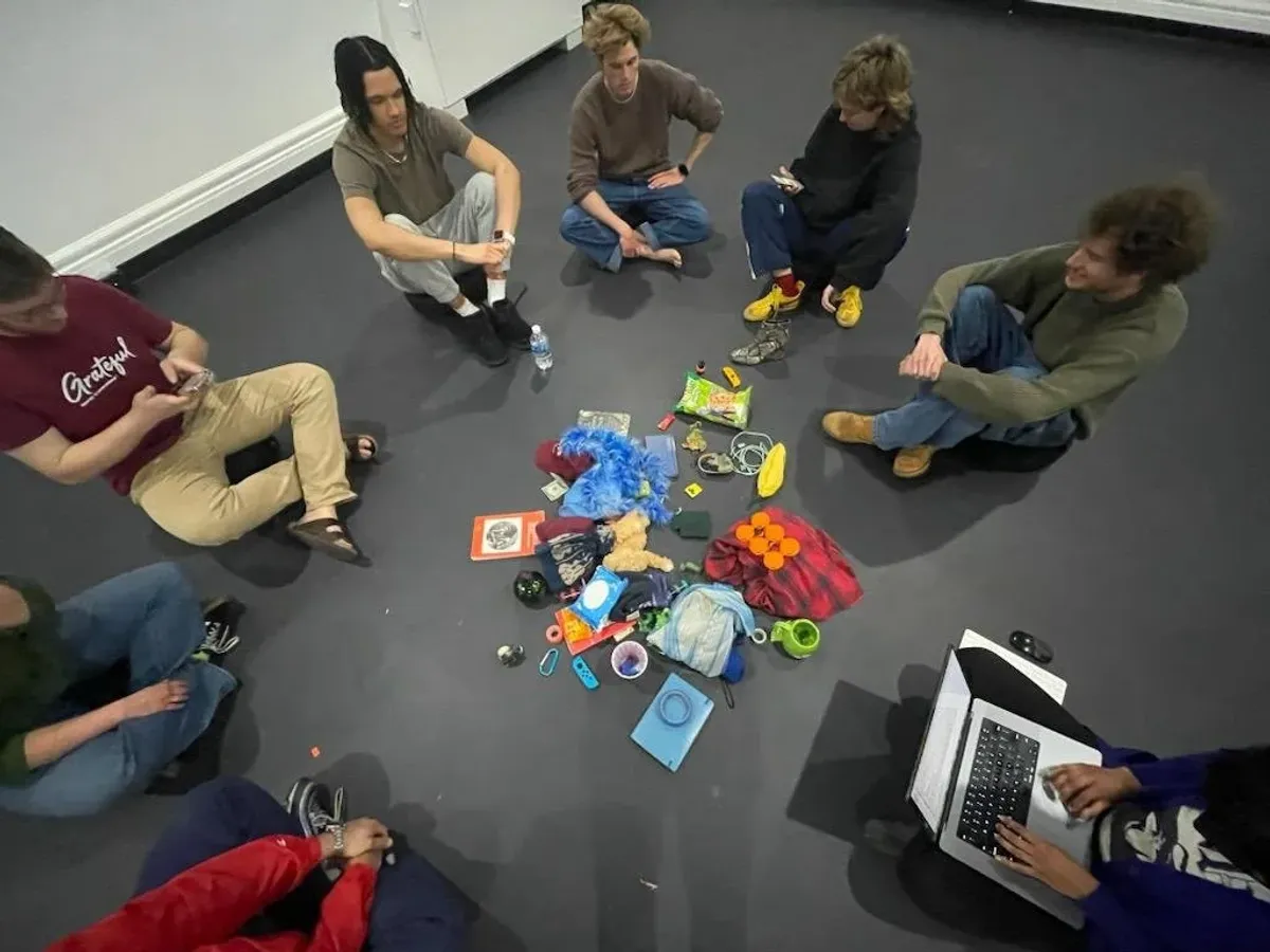 Wide overhead view of a circle of seated participants around the pile of collected personal items, one person typing on a laptop.