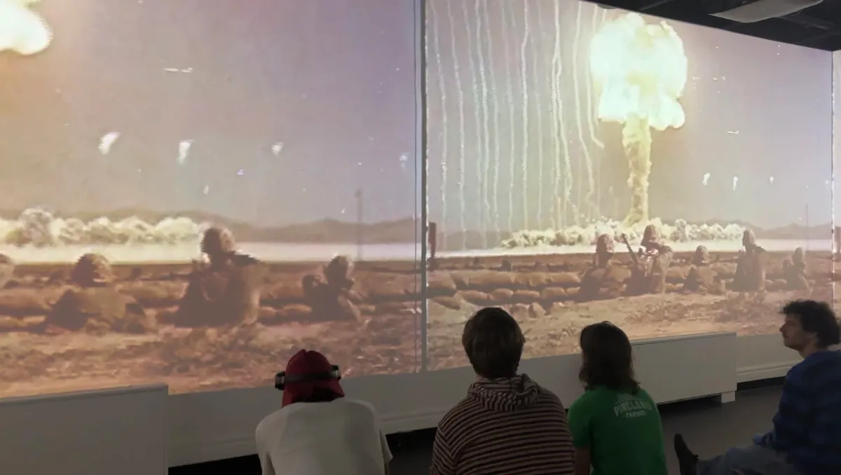 Installation view of archival atomic bomb test footage — a mushroom cloud rising over a desert landscape — projected across a wraparound screen, viewers seated below.