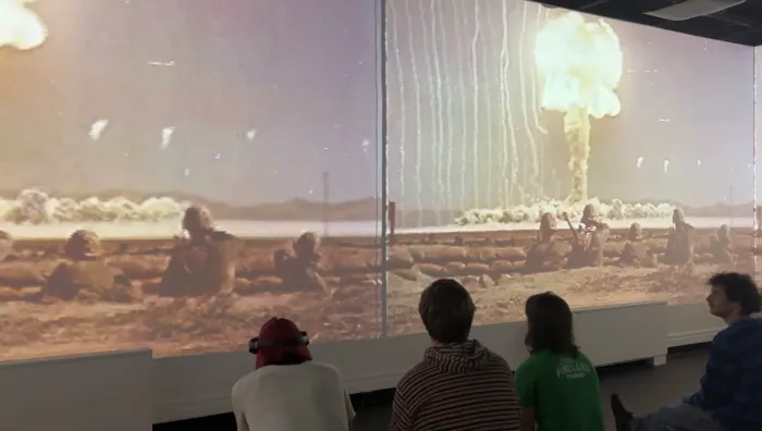 Installation view of archival atomic bomb test footage — a mushroom cloud rising over a desert landscape — projected across a wraparound screen, viewers seated below.
